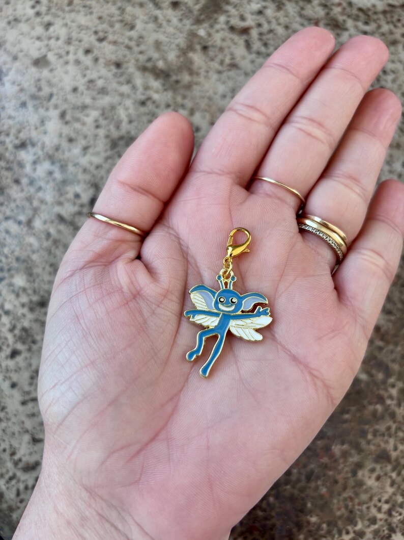 May include: A gold-coloured charm featuring a blue and white cartoon fairy design. The fairy has large eyes, a small mouth, and outstretched wings.