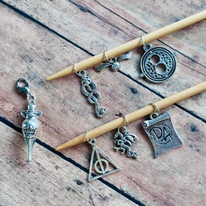May include: A set of eight silver charm stitch markers featuring Harry Potter themed designs, including a potion bottle, a snake, a golden snitch, a Deathly Hallows symbol, a dragon, a Time-Turner, and a DA badge. The charms are attached to two wooden knitting needles.