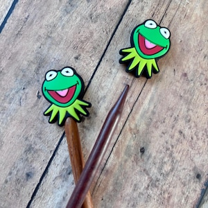 May include: Two wooden knitting needles with green frog-shaped stitch markers. The frog has a wide smile and large eyes.