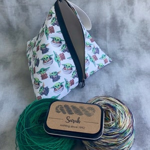 May include: A white zippered pouch with a repeating pattern of green baby Yoda characters. Two balls of yarn, one green and one multi-colored, are next to a small tin with a brown label that reads "Sarah knitting since 1992".
