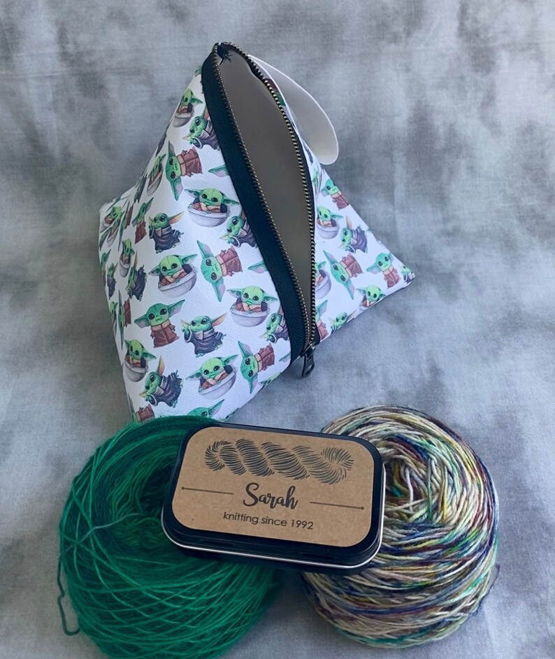 May include: A white zippered pouch with a repeating pattern of green baby Yoda characters. Two balls of yarn, one green and one multi-colored, are next to a small tin with a brown label that reads "Sarah knitting since 1992".