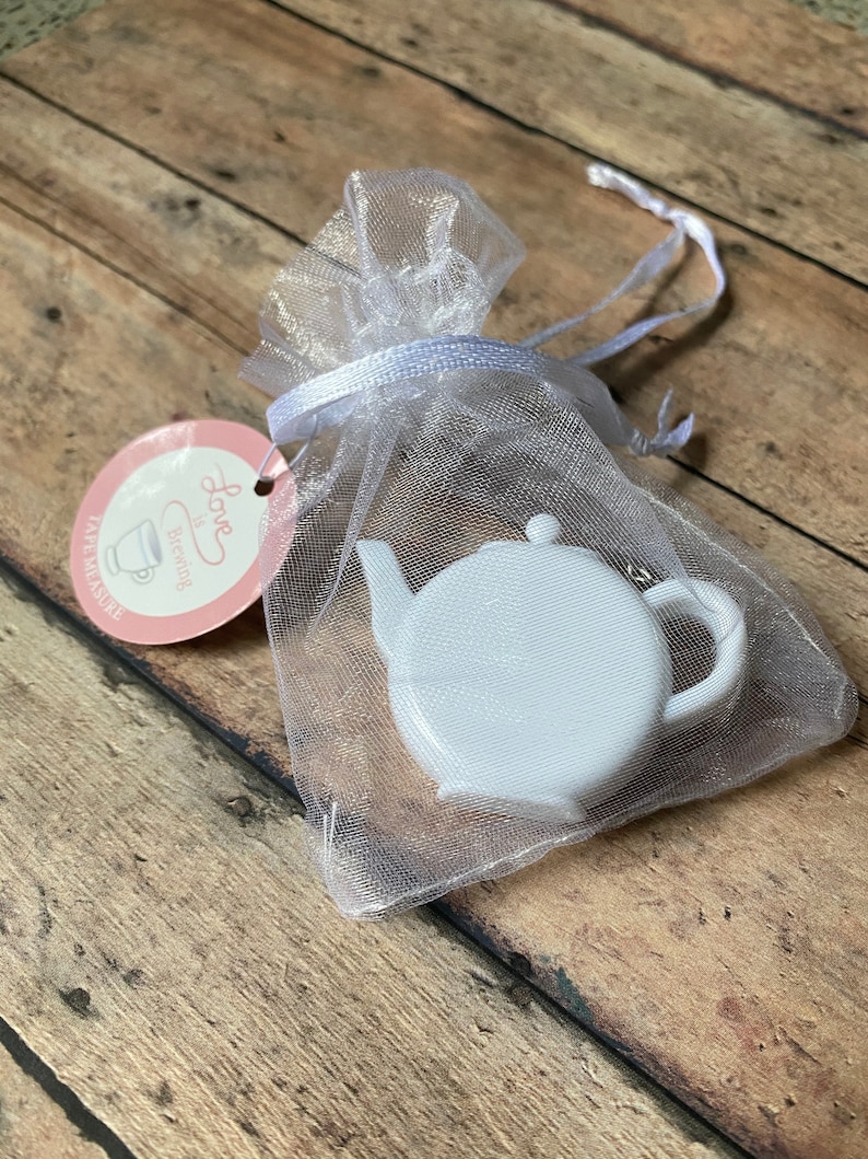 May include: A white ceramic teapot charm in a white organza bag with a pink tag that says "Love the Brew" and "Cafe Miniature".