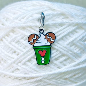 May include: A green and white enamel charm shaped like a coffee cup with a Mickey Mouse design. The charm features white whipped cream and two gingerbread cookie ears. A silver clasp is attached to the top.