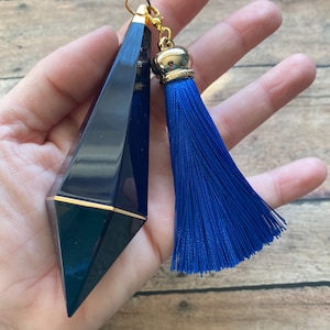 May include: A dark blue, faceted, glass bottle stopper with a gold top and a blue tassel. The bottle stopper is held in a hand.