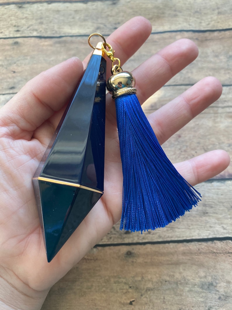 May include: A dark blue, faceted, glass bottle stopper with a gold top and a blue tassel. The bottle stopper is held in a hand.