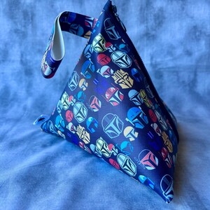 May include: A blue triangular pouch with a white handle. The pouch is covered in a repeating pattern of colorful Mandalorian helmets.