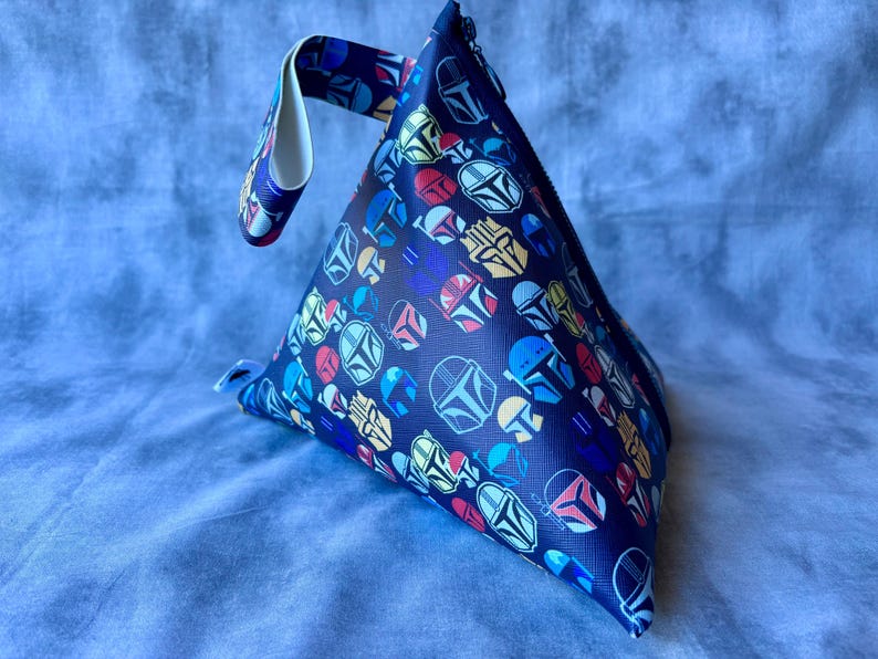 May include: A blue triangular pouch with a white handle. The pouch is covered in a repeating pattern of colorful Mandalorian helmets.