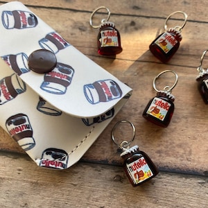 May include: A white zipper pouch with a Nutella pattern and six small Nutella-themed keychains. The keychains are red and brown with the Nutella logo and a silver ring.