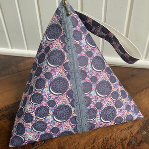 May include: A gray and white triangular zippered pouch with a black and white Oreo cookie print. The pouch has a white strap with a black and white Oreo cookie print. The pouch is on a wooden surface.
