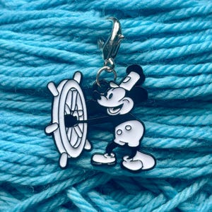 May include: A black and white Mickey Mouse charm holding a ship's wheel, attached to a silver-colored lobster clasp. The charm is set against a background of blue yarn, perfect for crafting projects.
