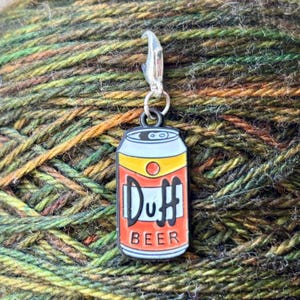 May include: A metal beer can charm with a red and yellow design. The can has the word "Duff" and "Beer" printed on it. The charm is attached to a silver metal lobster clasp.