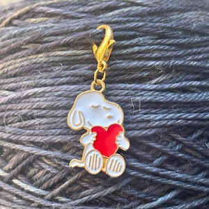 Op de afbeelding: Een goudkleurige bedel met een witte cartoonhond die een rood hart vasthoudt. De bedel heeft een karabijnsluiting voor bevestiging. De hond heeft zwarte ogen en een zwarte neus. De bedel is waarschijnlijk voor een armband of ketting.