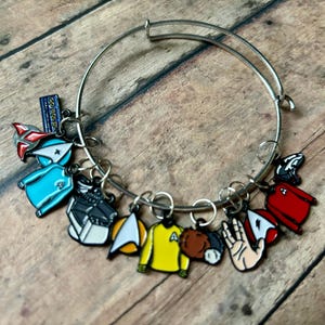 May include: Silver-tone bangle bracelet adorned with colorful enamel charms. Charms include a blue uniform, a yellow shirt, a Vulcan salute, and other Star Trek-themed designs. The bracelet is displayed on a wooden surface.
