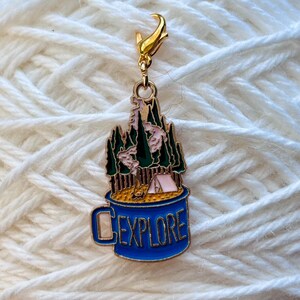 May include: A blue enamel mug with a campfire scene and the word "EXPLORE" on the side. The mug is attached to a gold metal lobster clasp.