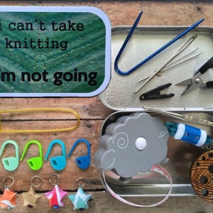 May include: A metal tin with a quote that reads "if I can't take my knitting I'm not going". The tin is filled with knitting supplies, including stitch markers, a tape measure, a yarn needle, and a pair of scissors.