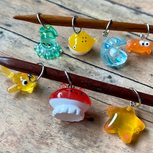 Puede incluir: Un conjunto de ocho marcadores de punto de colores con forma de criaturas marinas. Los marcadores están hechos de vidrio y presentan una variedad de criaturas marinas, incluyendo una medusa, una estrella de mar, una almeja, un pez globo, un cangrejo, un pulpo y un camarón. Los marcadores están unidos a dos agujas de tejer de madera.