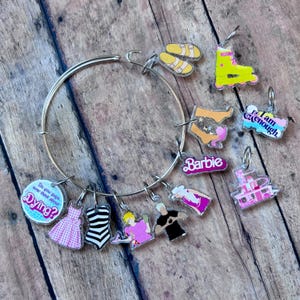 May include: A silver charm bracelet with various charms, including a pink and white gingham dress, a black and white striped swimsuit, a pink and white roller skate, a pink and white house, a pink and white Barbie logo, a pink and white "I am enough" charm, a pink and white "Do you guys ever think about dying?" charm, and a pink and white "Barbie" charm.