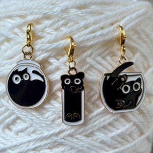 May include: Three gold cat-themed stitch markers. Each marker features a black cat in a different pose. One cat is curled up in a ball, another is standing on its hind legs, and the third is peeking out of a fishbowl.