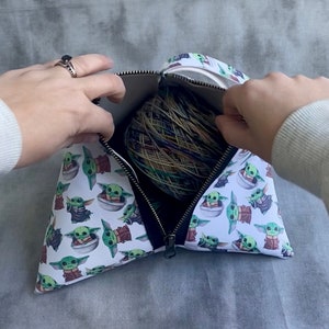 May include: A white zippered pouch with a black zipper and a repeating pattern of green baby Yoda characters. The pouch is open and a ball of yarn is inside.