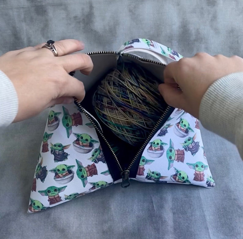 May include: A white zippered pouch with a black zipper and a repeating pattern of green baby Yoda characters. The pouch is open and a ball of yarn is inside.