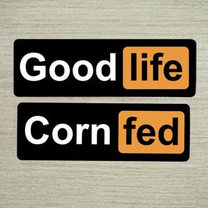 May include: Two black rectangular stickers with white text. The top sticker reads "Good life" with "life" in an orange box. The bottom sticker reads "Corn fed" with "fed" in an orange box.