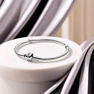 May include: A silver bracelet with a snake chain design. The bracelet has a square charm with engraved letters and a secure clasp. The jewellery is displayed on a white surface with a soft, neutral background.