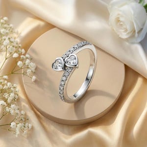May include: A silver-toned bypass ring featuring two heart-shaped clear stones at the open ends, with the band encrusted with a row of small, round, clear pavé stones along the shank, creating a continuous sparkling effect on the metal surface.