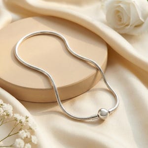 May include: A silver-toned snake chain necklace featuring a smooth, spherical bead charm at the centre. The necklace is constructed with a flexible, rounded link design that maintains a consistent thickness throughout the entire length of the chain.