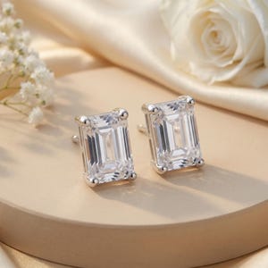 Può includere: Un paio di orecchini a lobo con pietre trasparenti taglio smeraldo incastonate in metallo color argento lucido. Ogni pietra rettangolare è fissata da quattro griffe e presenta un design sfaccettato che riflette la luce in modo brillante.
