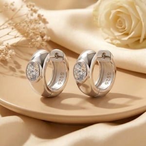 May include: A pair of silver hoop earrings featuring a polished finish and a single round clear stone set in the front. Each earring is engraved with the text S925 on the inner side, indicating the sterling silver composition of the jewelry.