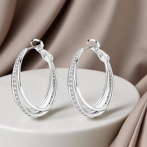 May include: A pair of silver hoop earrings with a double-band design. The outer band is encrusted with small, sparkling crystals. The earrings are displayed on a white surface against a soft, neutral-toned background.