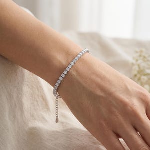 May include: A silver-toned tennis bracelet featuring a continuous row of round, clear, faceted stones set in a metallic frame. The bracelet includes an adjustable chain extension with a small, flat, teardrop-shaped metal charm at the end.