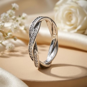 Può includere: Un anello color argento caratterizzato da un design a fascia intrecciata e incrociata. Un filo della fascia è incastonato con una fila continua di piccole pietre chiare a taglio rotondo, mentre l'altro filo ha una finitura metallica liscia e lucida.
