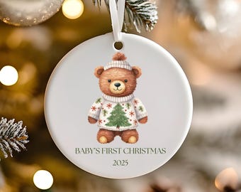 Puede incluir: Adorno navideño redondo de cerámica blanca con una ilustración de acuarela de un osito de peluche con un suéter navideño. El texto "BABY'S FIRST CHRISTMAS 2025" está impreso debajo. Se adjunta una cinta blanca para colgar.