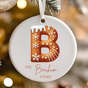 May include: A white ceramic Christmas ornament with a gingerbread-style letter "B" design. The letter is brown with white icing and snowflake details. The text "THE Beachum FAMILY" is written in a copper color.