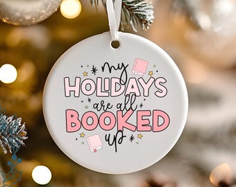Könnte beinhalten: Weißer Keramik-Weihnachtsschmuck mit dem Text "my HOLIDAYS are all BOOKED up" in Rosa und Schwarz. Der Schmuck hat ein Band zum Aufhängen und ist mit Sternen und Buchillustrationen verziert.