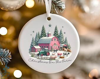 May include: A white ceramic Christmas ornament featuring a farm scene. A pink barn, tractor, and various farm animals are depicted. The text "Christmas On The Farm" is written in a script font. The ornament has a hole for hanging with a white ribbon.
