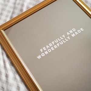 May include: A gold-coloured framed mirror with the text "FEARFULLY AND WONDERFULLY MADE" in white lettering. The mirror has a reflective surface and is set against a neutral-toned background. The frame has a detailed, ornate design.