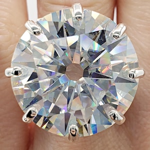 May include: A large, round, clear gemstone ring with a silver setting. The gemstone is faceted, catching and reflecting light. The ring is held by a hand, with the initials "B.B" in script at the bottom.