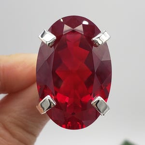 May include: A large oval-cut deep red gemstone set in a silver-toned metal ring. The stone features multifaceted surfaces that reflect light, secured by four prominent prongs at the corners. The letters BB are visible in white at the bottom.