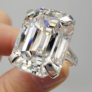 May include: A large, rectangular-cut diamond ring with a silver band. The diamond is clear and faceted, reflecting light. The ring has a classic design with four prongs securing the stone. The initials "BB" are at the bottom of the image.