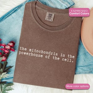 May include: Espresso-coloured t-shirt with the text "the mitochondria is the powerhouse of the cell." The shirt is folded on top of blue jeans. The shirt has a Comfort Colors tag.