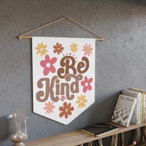 May include: A white wall hanging with the words "Always Be Kind" in brown and pink lettering, surrounded by floral designs in pink, yellow, and brown. The banner is suspended from a wooden rod with a natural cord.