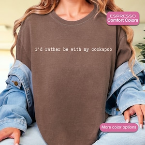 May include: A brown comfort colors t-shirt with the text "i'd rather be with my cockapoo" in white. The shirt is worn with a denim jacket. The image also includes a pink rectangle with the words "More color options".