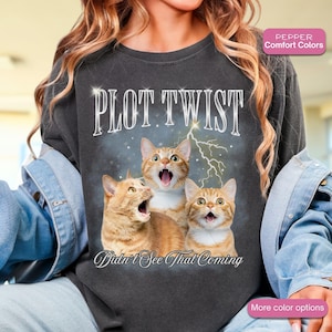 May include: Dark gray sweatshirt featuring three surprised orange tabby cats with the text "PLOT TWIST" and "Didn't See That Coming." The design includes a lightning bolt and stars, creating a humorous and eye-catching graphic.