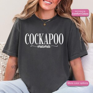 May include: A dark gray t-shirt with the words "COCKAPOO mama" in white script. The shirt is a comfort color and has a relaxed fit. The image also includes a pink rectangle with the words "More color options".