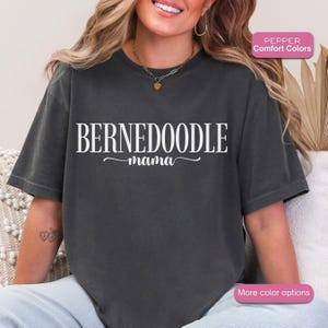 May include: Dark gray t-shirt with the words "BERNEDOODLE mama" in white script. The shirt is a comfort color and has a relaxed fit. The image also includes a small gold necklace.