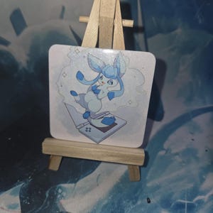 Op de afbeelding: Vierkante, lichtblauwe illustratie van een stripfiguur met lange oren en een blauw-wit kleurenschema, staand op een draagbaar spelapparaat. Het kunstwerk wordt gepresenteerd op een klein houten schildersezel.