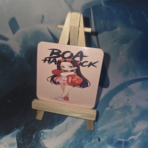 Può includere: Una stampa artistica quadrata, rosa e glitterata con un'illustrazione di un personaggio dei cartoni animati con lunghi capelli neri, che indossa un abito rosso, e la scritta "BOA HANCk". La stampa è esposta su un piccolo cavalletto di legno.