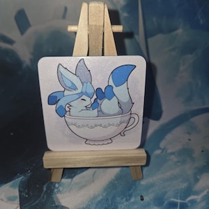 Op de afbeelding: Een vierkante onderzetter met een cartoon illustratie van een blauw vosachtig wezen dat ontspant in een witte theekop. De onderzetter wordt gepresenteerd op een klein houten ezel.